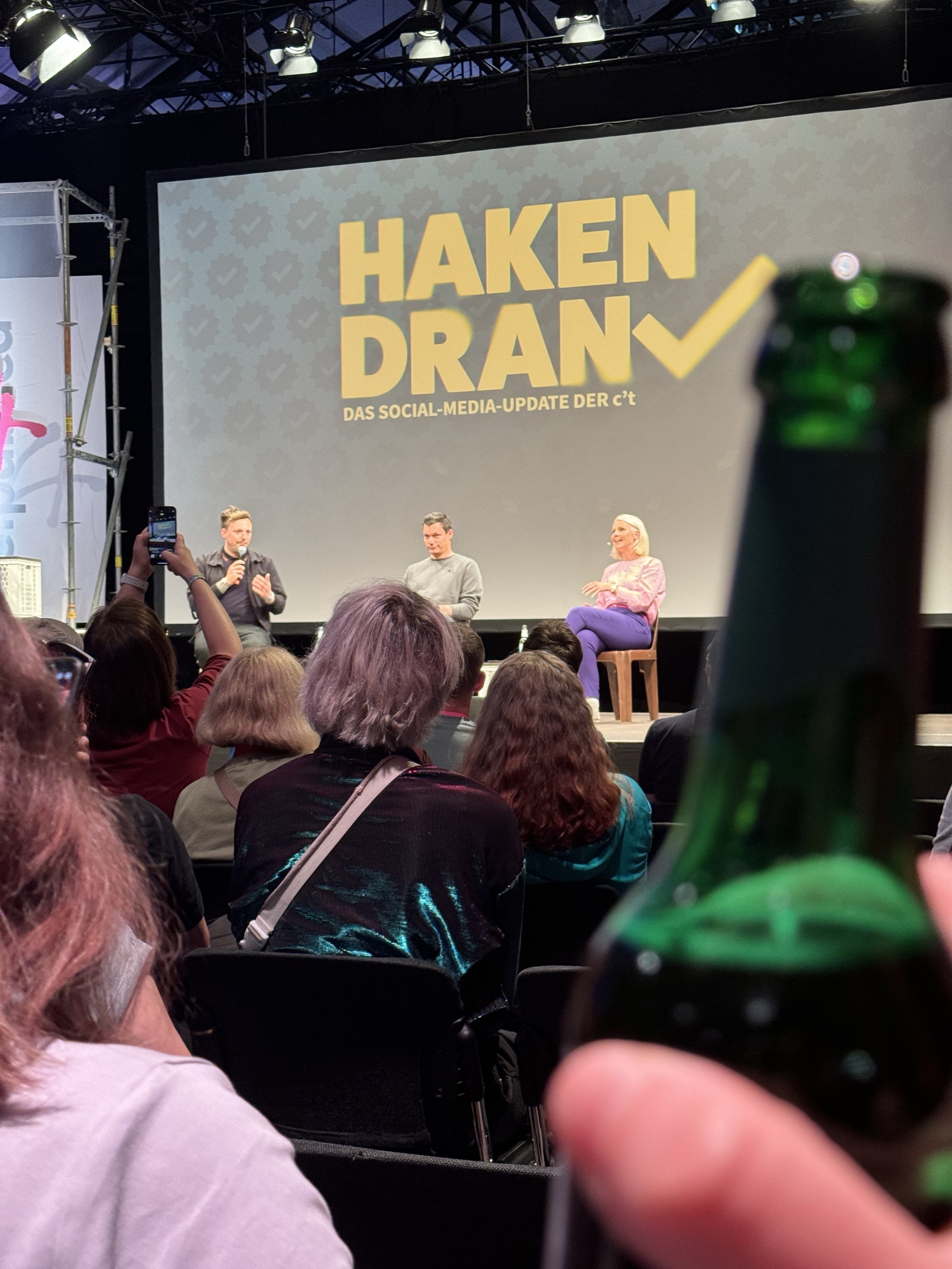 Podiumsdiskussion auf der Bühne mit drei Rednern, einem Publikum und dem Text "HAKEN DRAN" im Hintergrund. Im Vordergrund unscharf eine Bierflasche.